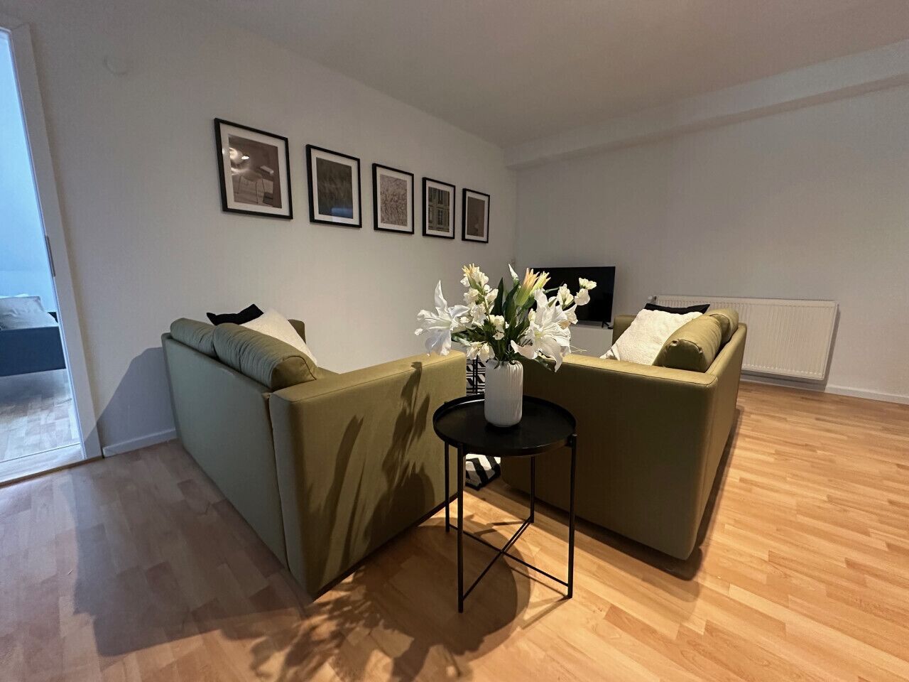 Una sala de estar con dos sofás verdes, una mesa de centro negra y un jarrón con flores. Hay cuatro cuadros en la pared.