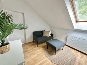 Ein modernes Schlafzimmer im Loft-Stil mit schrägem Dach, einem dunkelgrauen Sofa, einem kleinen grauen Hocker, einer Topfpflanze und Blick ins Grüne durch ein Dachfenster.