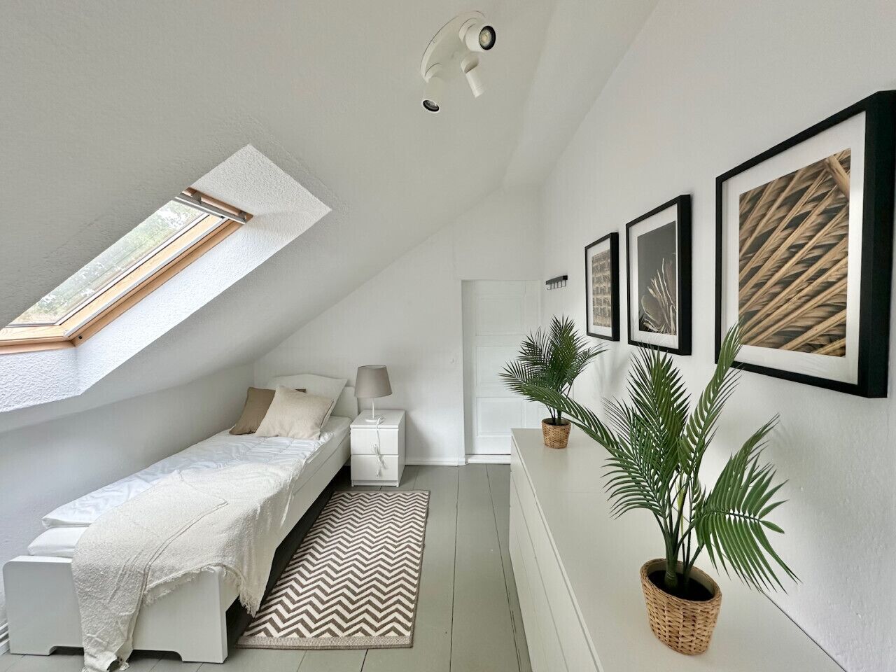 Une chambre à coucher avec un puits de lumière, un lit, une table de chevet, une plante en pot et des tableaux encadrés accrochés au mur.