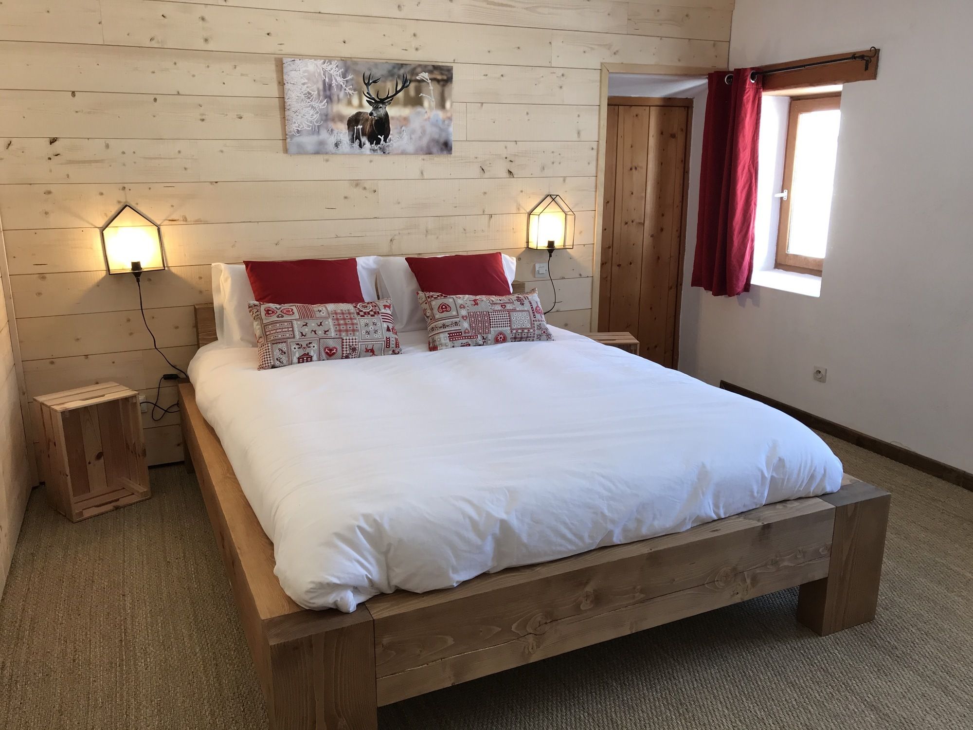 A wooden bed with white bedding, two bedside lamps, a red curtain, and a wall-mounted painting of a deer.