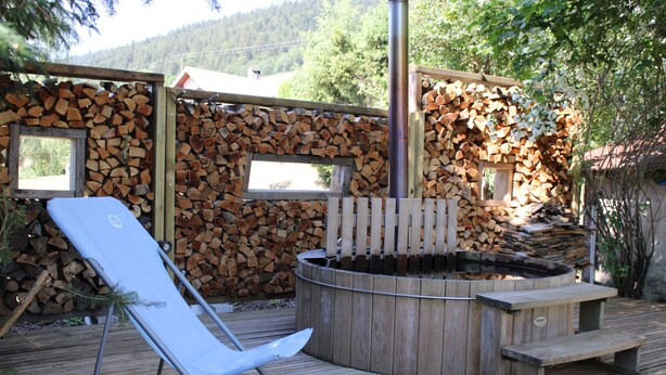 A wooden deck with a hot tub, a lounge chair, and a stack of firewood.
