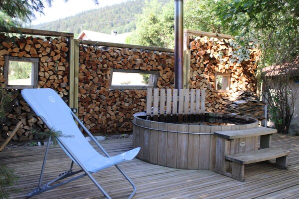 A wooden deck with a hot tub, a lounge chair, and a stack of firewood.