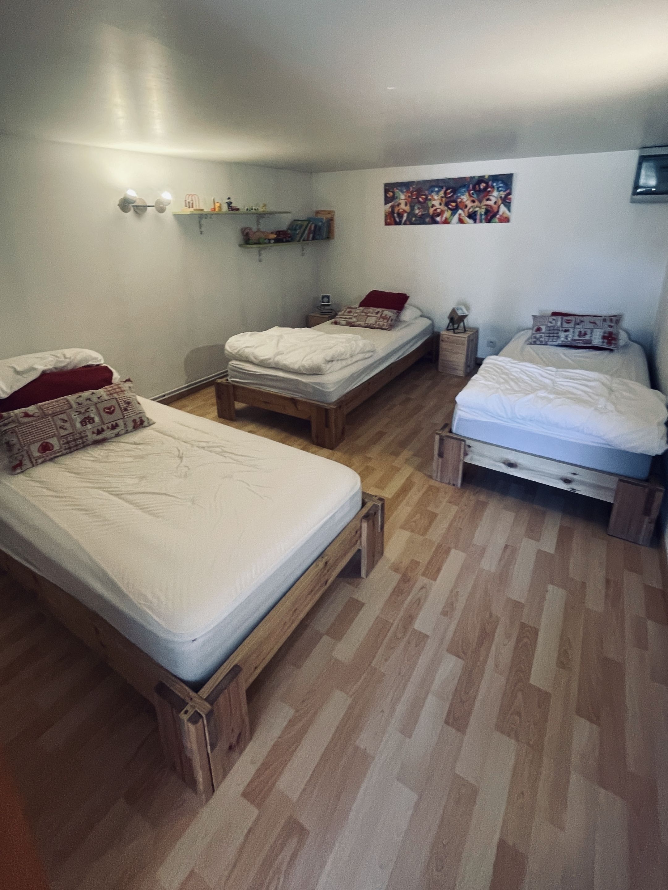 Een kamer met drie houten bedden, een plank met boeken en kleine spullen, en een kleurrijk schilderij aan de muur.