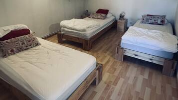 Een kamer met drie houten bedden, een plank met boeken en kleine spullen, en een kleurrijk schilderij aan de muur.
