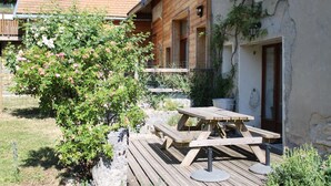 A wooden deck with a picnic table and benches, surrounded by greenery and a building with a balcony. - Le Corps de Ferme with Nordic bath (Autrans-Méaudre en Vercors)