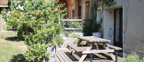 Une terrasse en bois avec une table de pique-nique et des bancs, entourée de verdure et bordée d’un bâtiment doté d’un balcon.