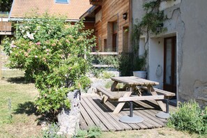 A wooden deck with a picnic table and benches, surrounded by greenery and a building with a balcony.
