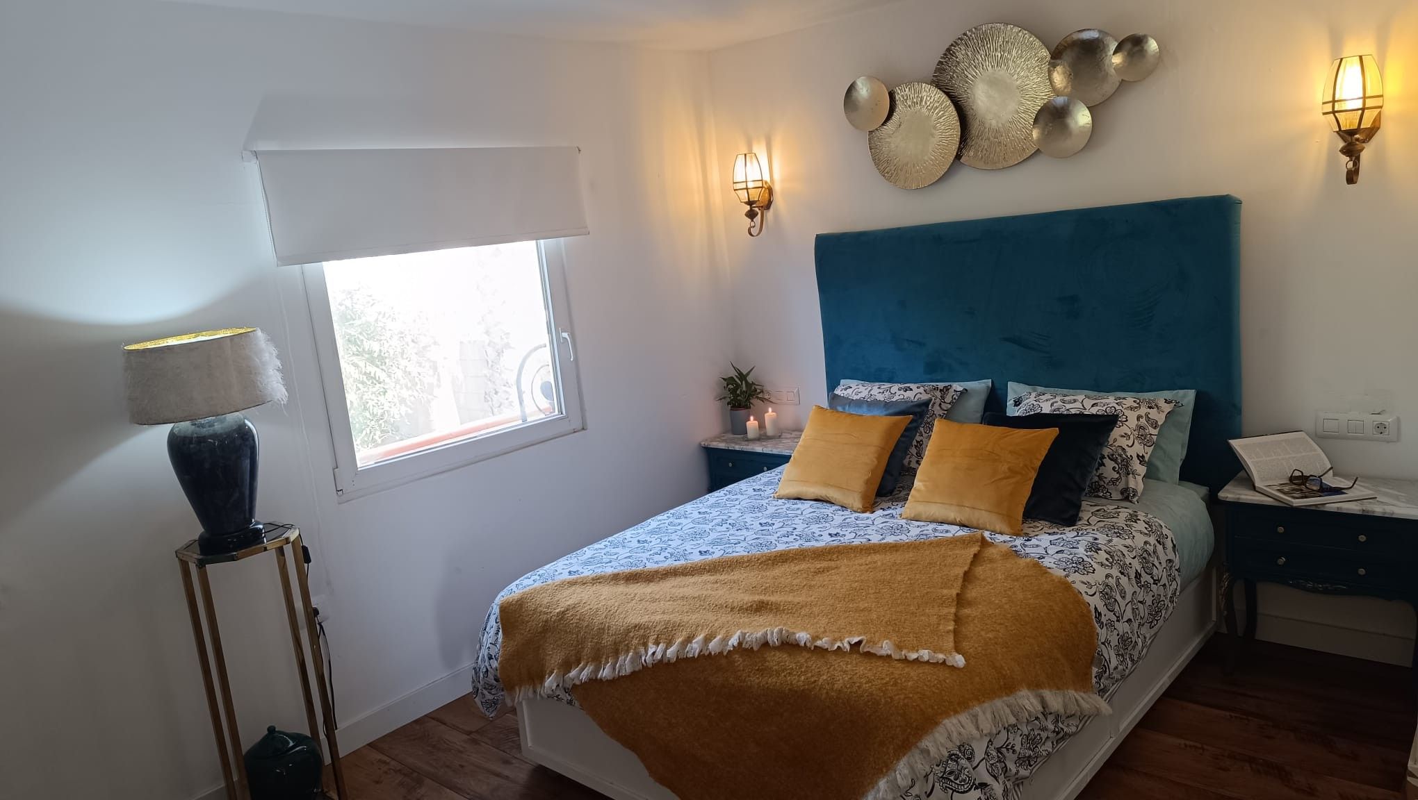Un dormitorio con cama, mesitas de noche, una lámpara, apliques de pared y una ventana.