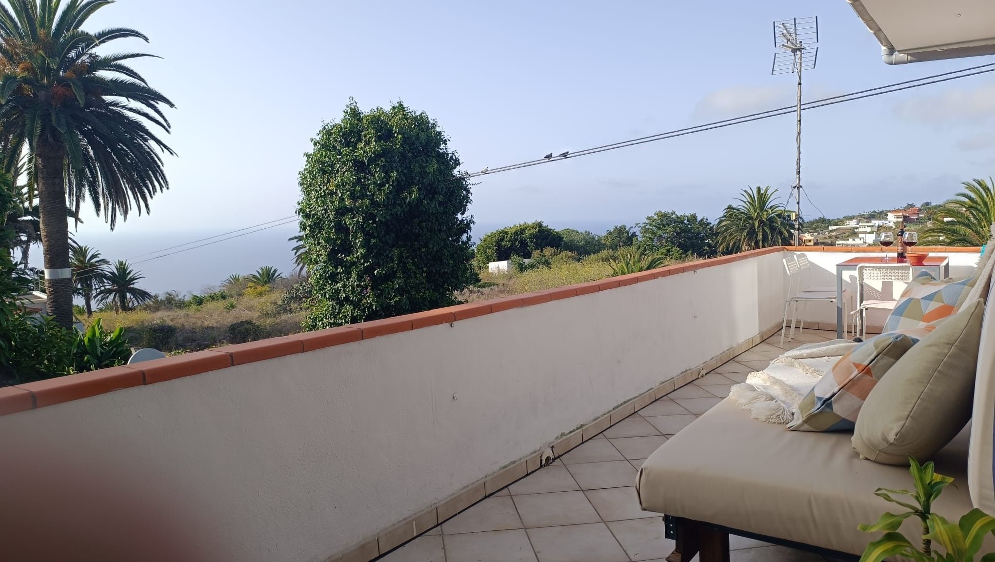 Un balcón con sofá, una mesa con vasos y vistas al mar y la vegetación.