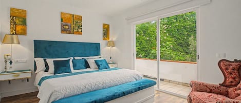 A bedroom with a large bed, two bedside tables, a chair, and a view of greenery through a sliding door.