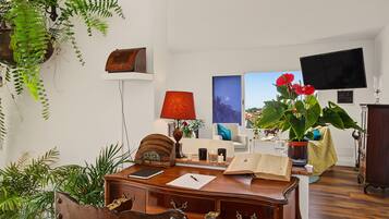 Una habitación con escritorio, libros, lámpara, televisor y varias plantas.