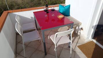 Un balcón con una mesa roja, dos sillas blancas, una botella de vino, dos copas de vino y un cojín azul.