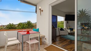 Ein Balkon mit Tisch und Stühlen, eine Glasschiebetür, die zu einem Wohnbereich mit Sofa und Fernseher führt.