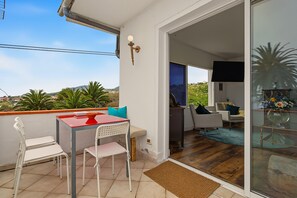 Un balcón con mesa y sillas, una puerta corrediza de vidrio que da paso a un espacio interior con sofá y televisor.