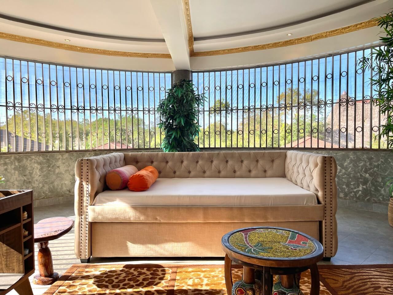A sunlit balcony with a tufted sofa, a round table, and a view of the outdoors.