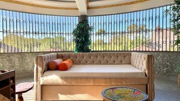 A sunlit balcony with a tufted sofa, a round table, and a view of the outdoors.