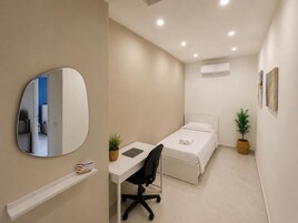 Una camera compatta con un letto singolo, una scrivania, una sedia, una pianta sul comodino e uno specchio fissato al muro.