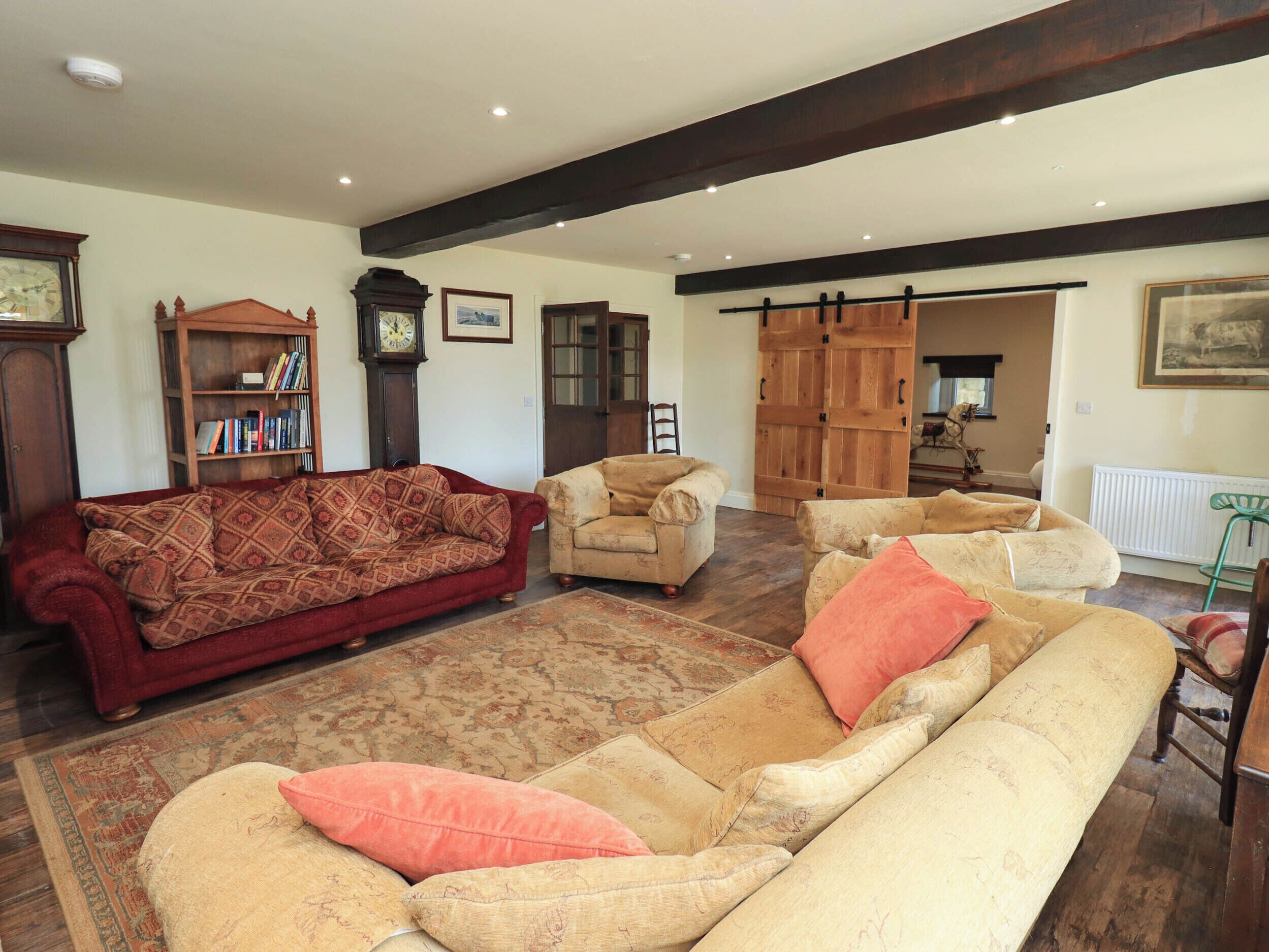 A living room with a red sofa, beige armchairs, a patterned rug, wooden bookshelves, a clock, and a wooden wardrobe.