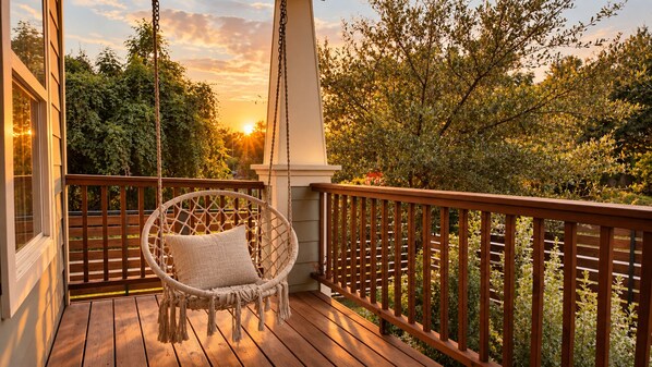 A wooden deck with a hanging chair and a view of trees and a sunset.