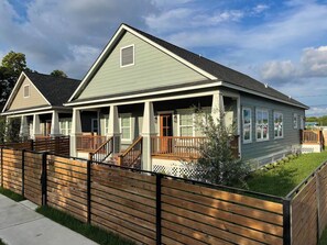 A two-story residential building with a covered porch, a fenced yard, and a clear sky.