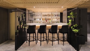 Una zona de bar con techo de madera, sillas negras y una estantería llena de botellas.