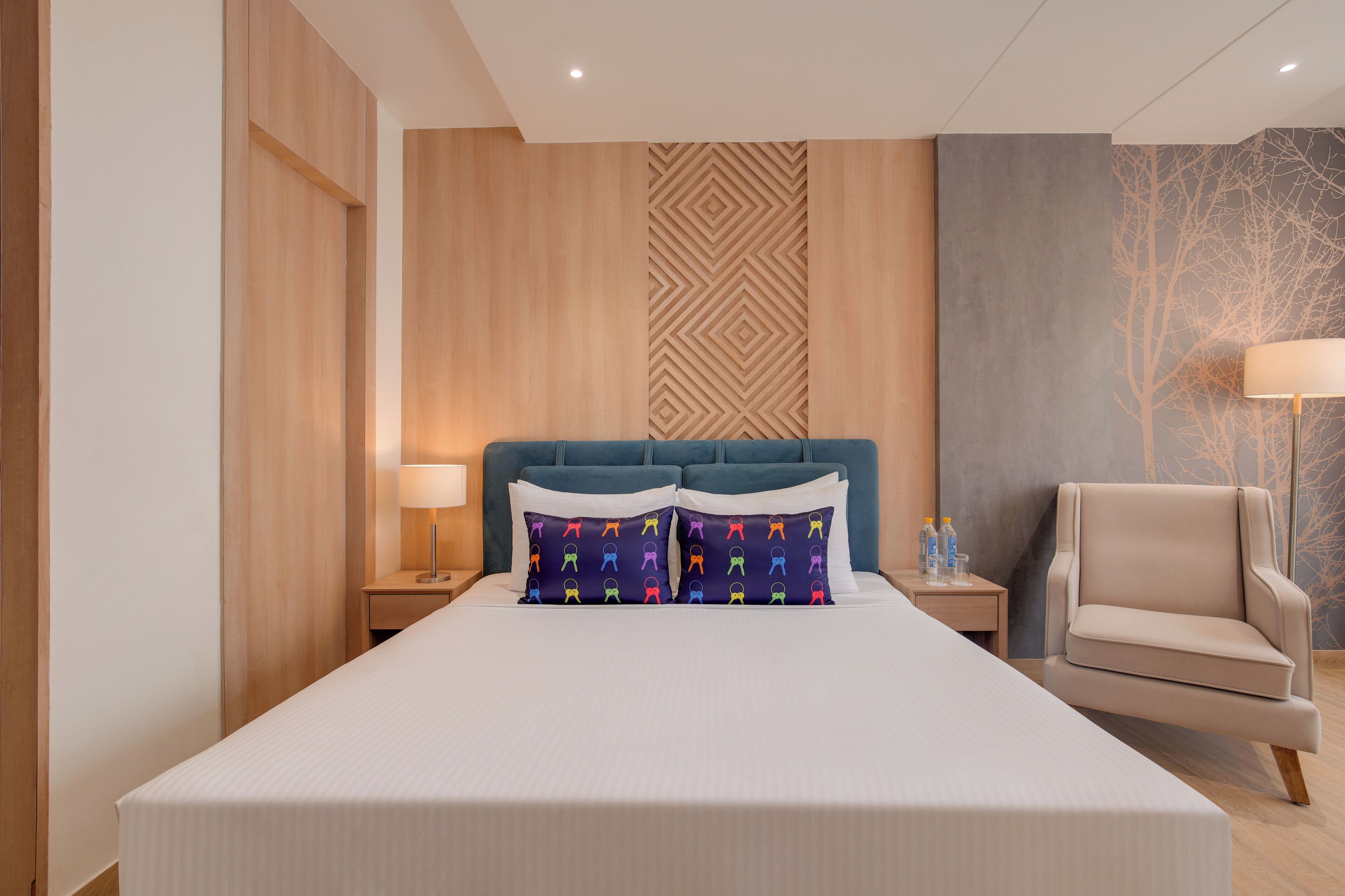 Habitación de hotel con una cama grande, cabecera, mesita de noche, una silla y una pared con diseño geométrico.