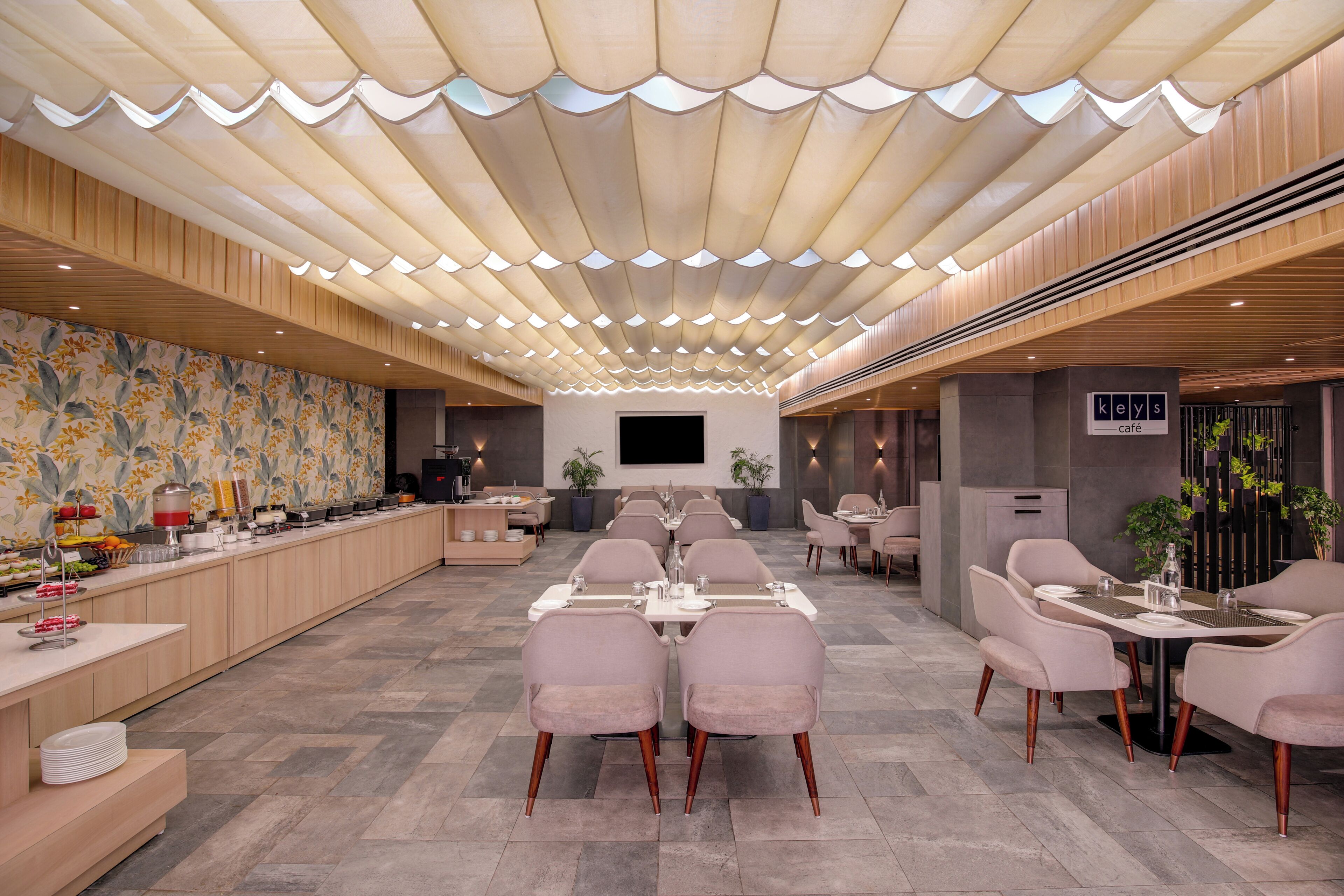 Amplia sala de comedor de hotel con varias mesas y sillas, un mostrador de buffet y un televisor de pantalla plana grande montado en la pared.