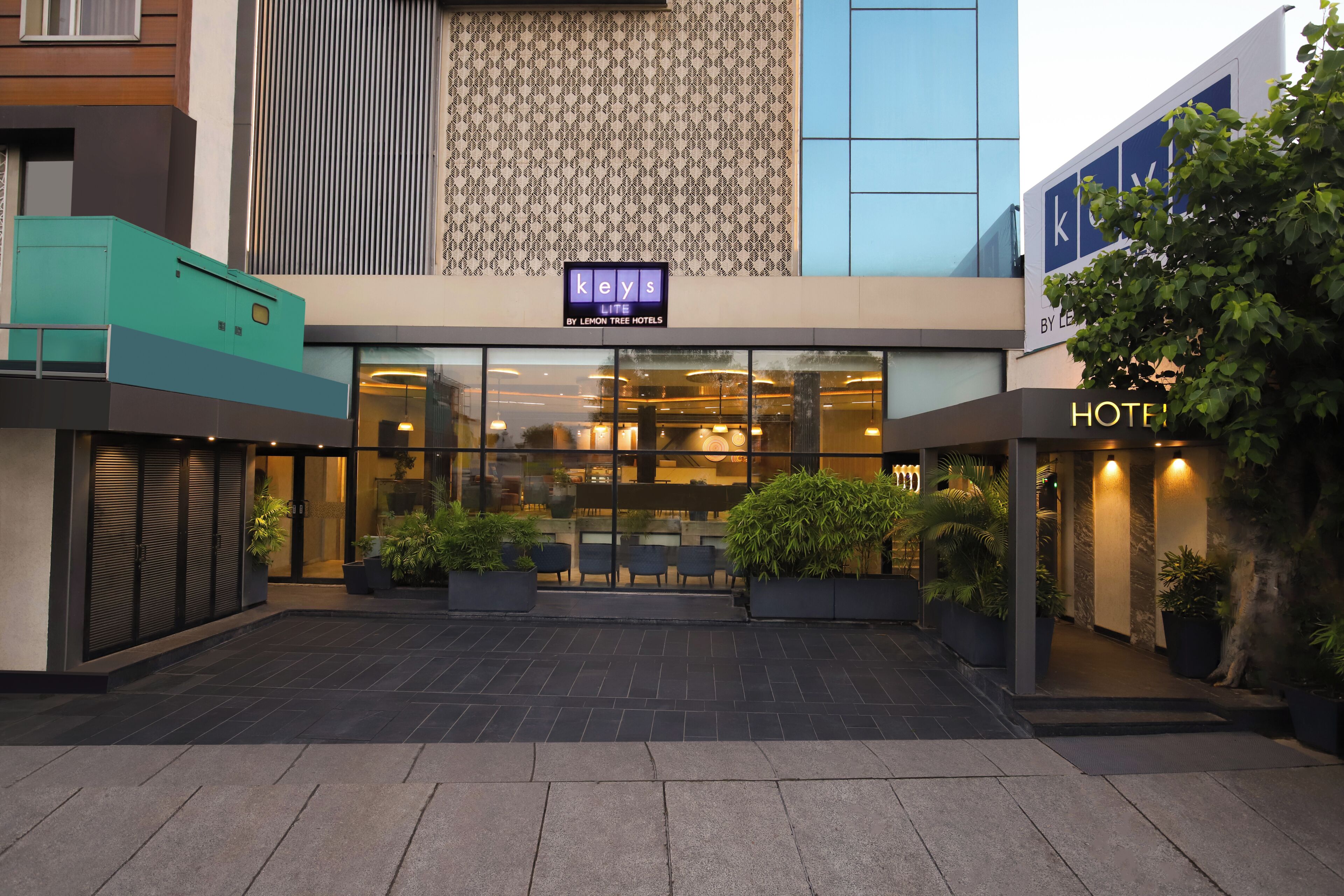 Entrada de hotel con un letrero que dice 'keys by lemon tree hotels', fachada de vidrio y plantas en macetas.