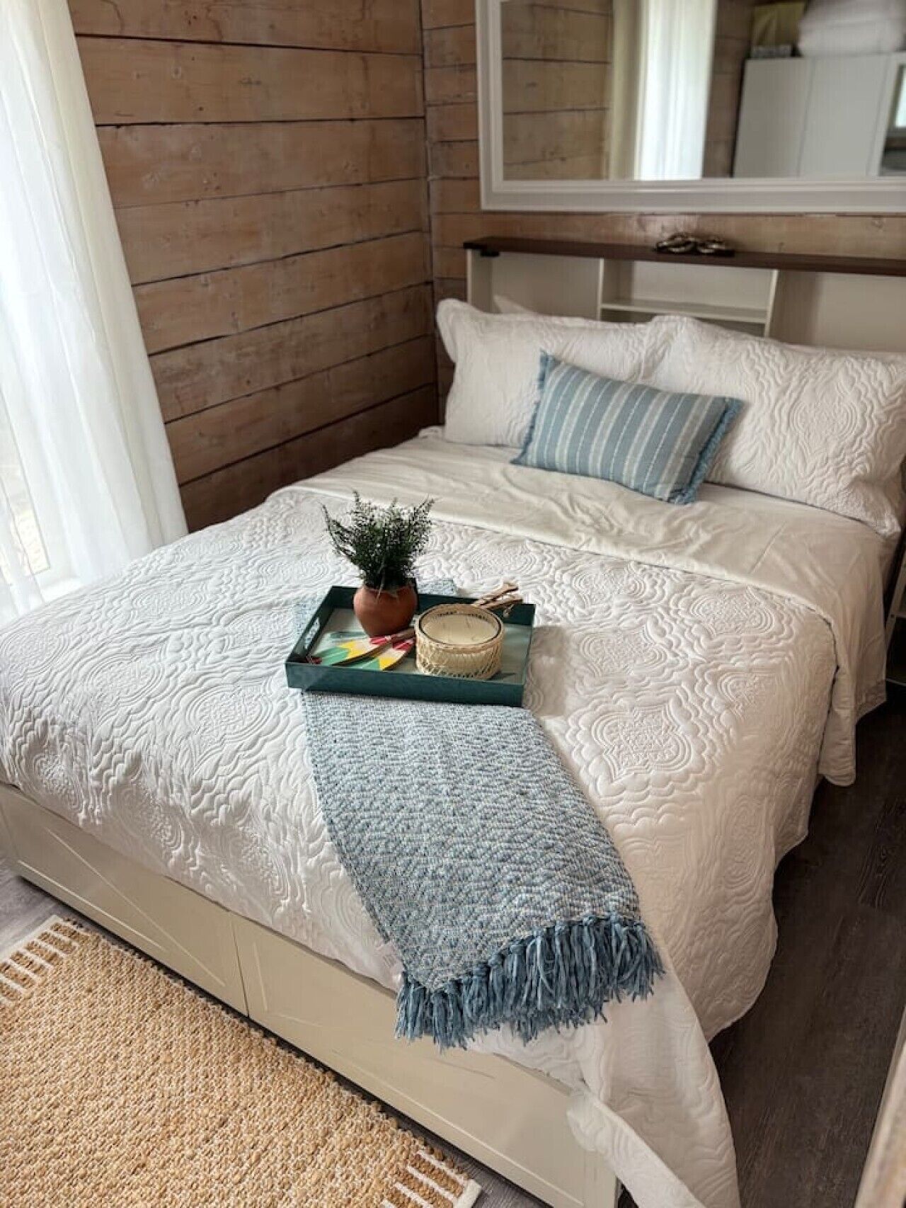 Un lit avec une couette blanche, un plaid bleu et un plateau sur lequel se trouvent une plante et une bougie.