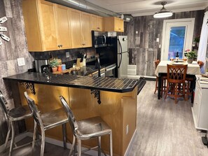 Une cuisine avec des meubles de rangement en bois, un plan de travail noir, des chaises en acier inoxydable, une table à manger avec des chaises et un réfrigérateur.