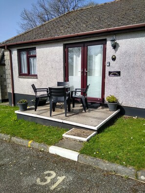 A single-story building with a tiled roof, a front door with glass panels, and a small outdoor seating area with two chairs and a table. - Tregenza self catering Holiday Bungalow, set in the grounds of 17cKenegie Manor. (Penzance)