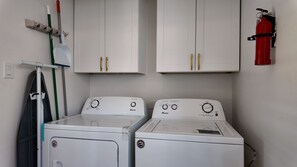 A laundry area with two white front-loading washers and dryers, a fire extinguisher on the wall, and a mop and broom leaning against the wall. - Houston 2BR Stay | Near NRG, & Downtown | Coffee (Houston)