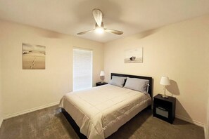 A bedroom with a bed, a ceiling fan, a nightstand with a lamp, a window with blinds, and two wall-mounted pictures. - Spring Branch 2BR Private Patio + Workspace (Houston)