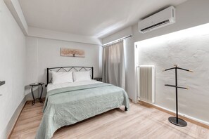 A bedroom with a bed, a bedside table, an air conditioning unit, a radiator, and a window with curtains. - Charming 1-Bedroom Garden Apartment Near Glyfada Center & Beach (Glyfada)