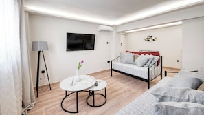 A modern living room with a sofa, a small round table, a TV, and an air conditioning unit. - Charming 1-Bedroom Garden Apartment Near Glyfada Center & Beach (Glyfada)