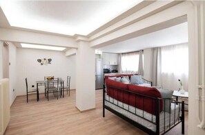 A compact living space with a dining area, a bed with red bedding, and a kitchenette in the background. - Charming 1-Bedroom Garden Apartment Near Glyfada Center & Beach (Glyfada)