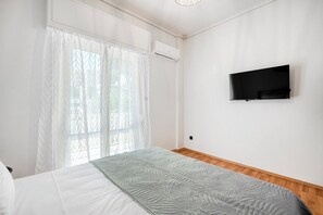 Quarto com cama, TV fixada na parede e janela com cortinas leves.