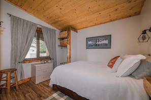 A bedroom with a wooden ceiling, a bed with pillows, a dresser, a window with curtains, and a picture on the wall. - The Old West Cabin Steps from the Lake (Sylvan Lake)