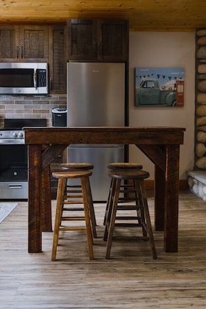 Uma cozinha com uma mesa de madeira e banquetas, uma geladeira, um micro-ondas e um quadro na parede.