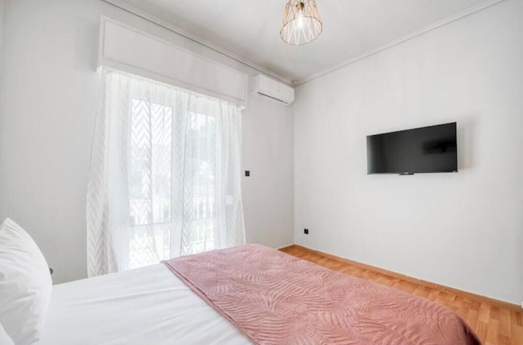 Une chambre à coucher avec un lit, un téléviseur fixé au mur et une fenêtre ornée de rideaux légers.