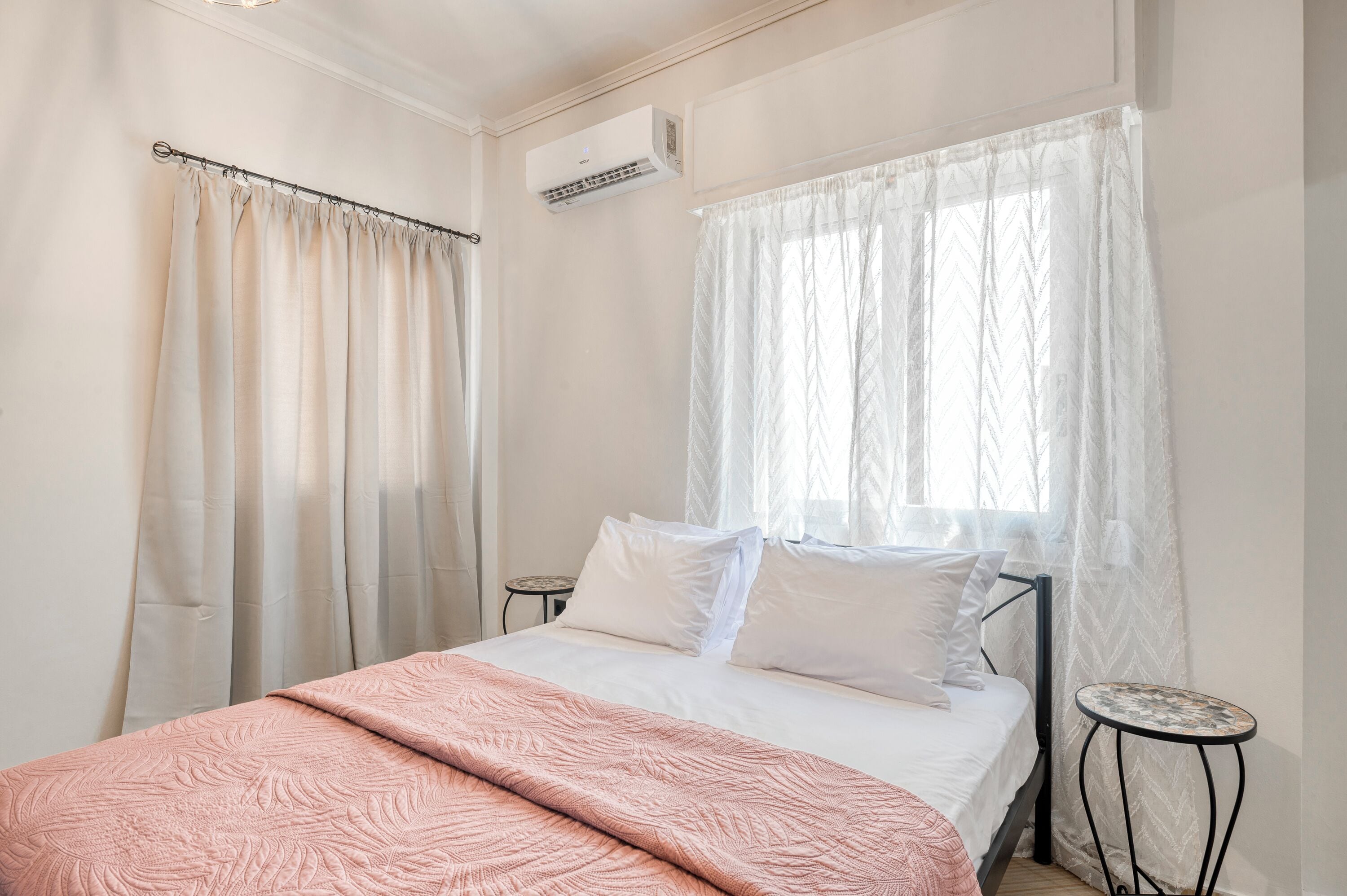 A bedroom with a bed, a bedside table, a window with sheer curtains, and an air conditioning unit.