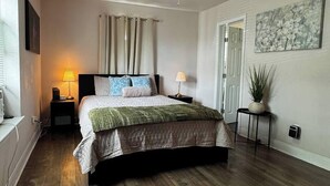 Un dormitorio con una cama, dos mesitas de noche, un ventilador de techo, una ventana con cortinas, una puerta y una planta colgada en la pared.