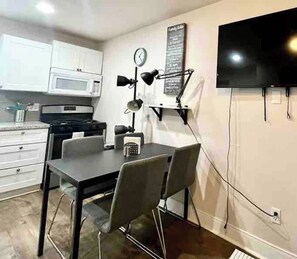 A compact kitchen with white cabinets, a microwave, a dining table with chairs, a clock, and a flat-screen TV mounted on the wall. - Restful Retreat w/Smart TV & Free Parking near NRG (Houston)