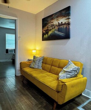 A yellow sofa with patterned pillows, a painting of a cityscape, and a lamp on a bedside table. - Restful Retreat w/Smart TV & Free Parking near NRG (Houston)