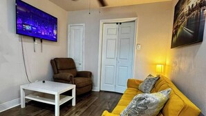 Una sala de estar con ventilador de techo, televisor montado en la pared, un sofá amarillo, un sillón marrón y una mesa de centro blanca.