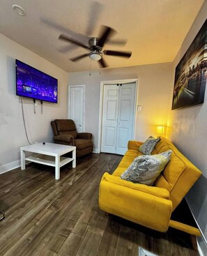 Ein Wohnzimmer mit Deckenventilator, einem an der Wand befestigten Fernseher, einem gelben Sofa, einem braunen Sessel und einem weißen Couchtisch.