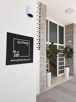 A hotel entrance with a signboard for 'GLYFADA STAY' and a website URL. There are two potted plants and a modern architectural design with a stone facade. - Loft Studio with Sea View | Modern Comfort Near Glyfada Beach & City Center (Glyfada)