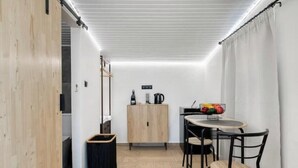 Une pièce de taille modeste, avec un plancher en bois, une petite table entourée de chaises, un meuble et une poubelle.