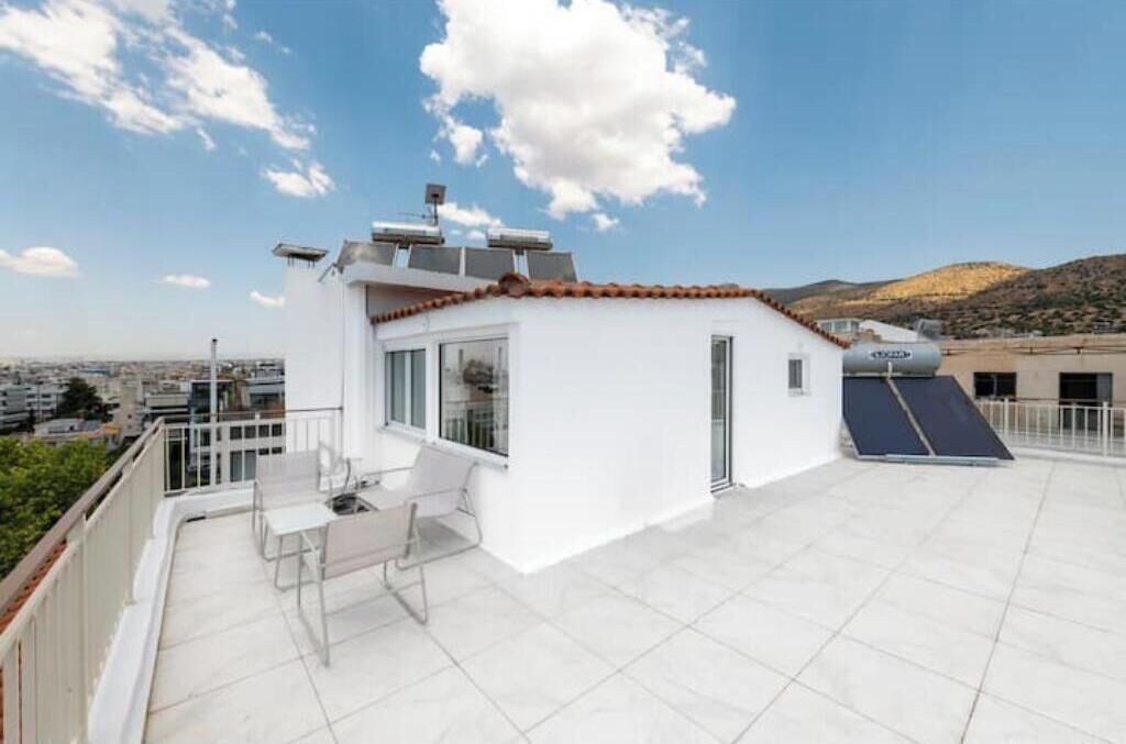 Eine Dachterrasse mit einem weißen Gebäude, einem Tisch mit Stühlen, einem Wassertank und Blick auf Stadt und Hügel.