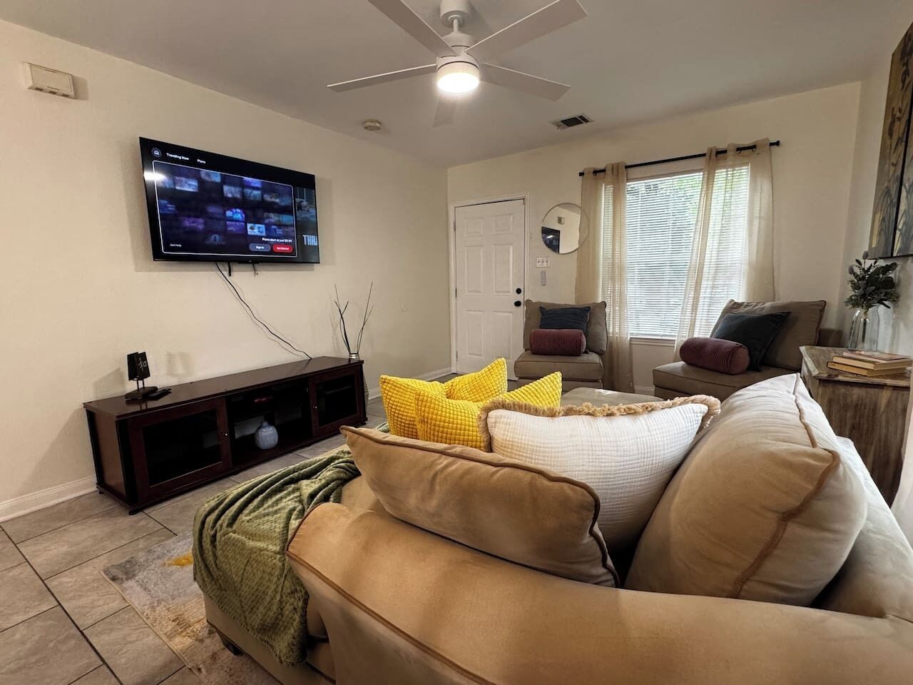 Una sala de estar con un televisor montado en la pared, un mueble de entretenimiento de madera oscura, un sofá beige con cojines amarillos y blancos, un sillón marrón y una ventana con cortinas.